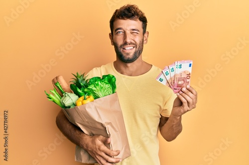 Handsome man with beard holding groceries and 10 euros banknotes winking looking at the camera with sexy expression, cheerful and happy face. photo