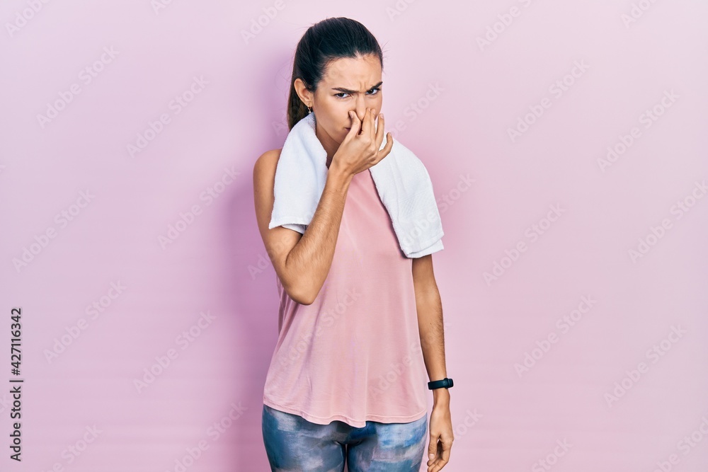 Young brunette woman wearing sportswear and towel smelling something stinky and disgusting, intolerable smell, holding breath with fingers on nose. bad smell