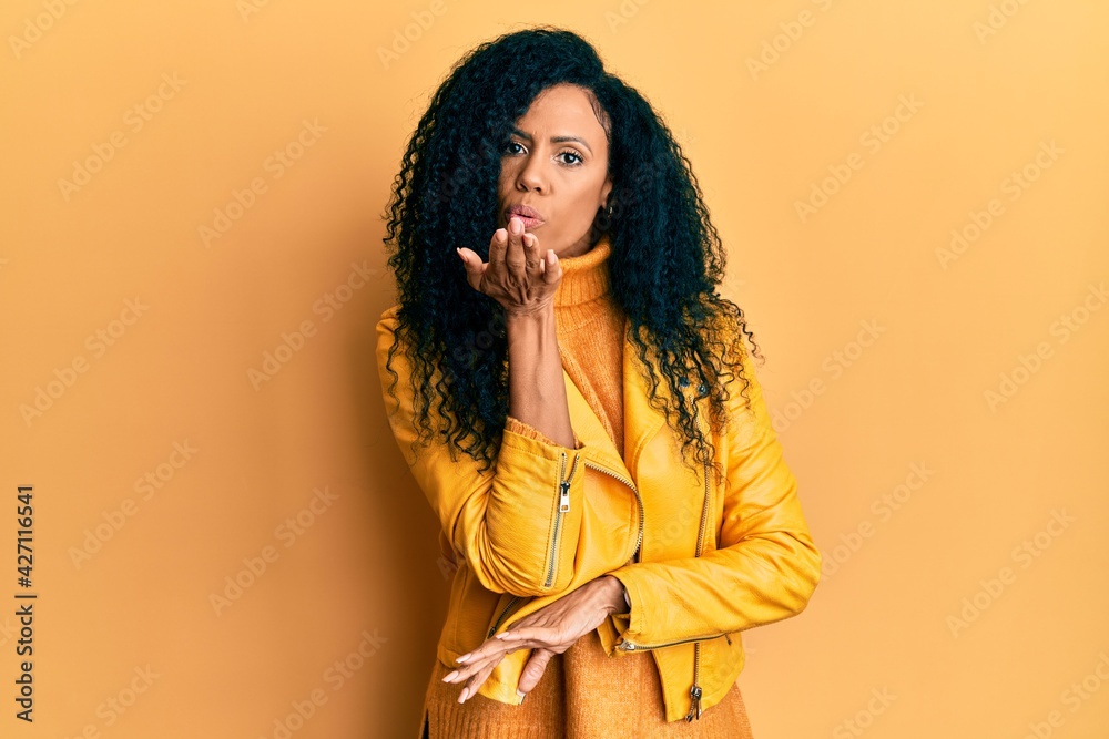 Middle age african american woman wearing wool winter sweater and leather jacket looking at the camera blowing a kiss with hand on air being lovely and sexy. love expression. Middle age african american woman wearing wool winter sweater and leather jacket looking at the camera blowing a kiss with hand on air being lovely and sexy. love expression.