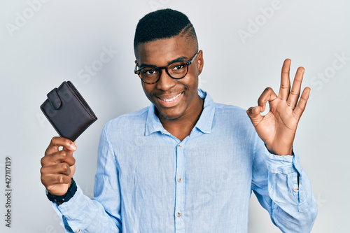 Young african american man wearing glasses holding leather wallet doing ok sign with fingers, smiling friendly gesturing excellent symbol © Krakenimages.com