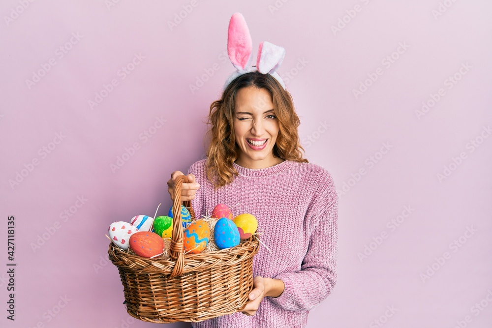 Young caucasian woman wearing cute easter bunny ears holding colored egg winking looking at the camera with sexy expression, cheerful and happy face.