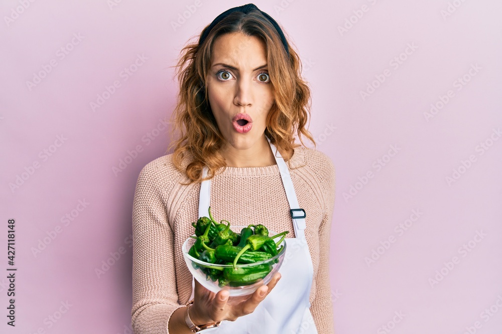 Young caucasian woman wearing apron holding green peppers bowl scared and amazed with open mouth for surprise, disbelief face Young caucasian woman wearing apron holding green peppers bowl scared and amazed with open mouth for surprise, disbelief face