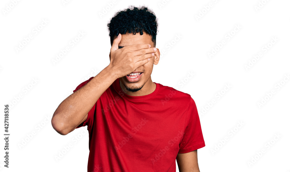 Young african american man wearing casual red t shirt smiling and laughing with hand on face covering eyes for surprise. blind concept. Young african american man wearing casual red t shirt smiling and laughing with hand on face covering eyes for surprise. blind concept.