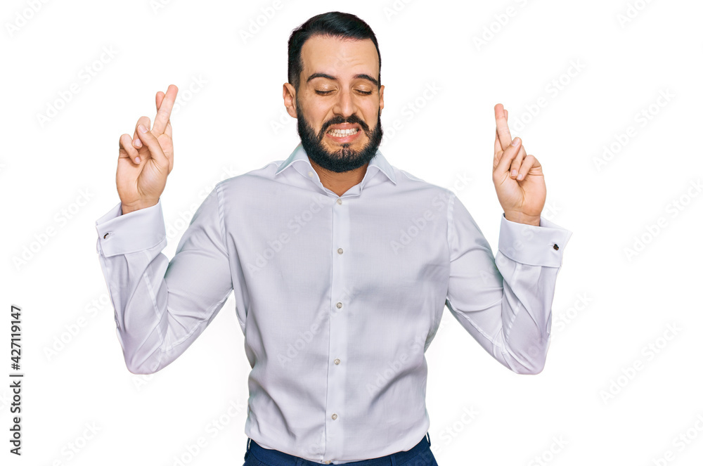 Young man with beard wearing business shirt gesturing finger crossed smiling with hope and eyes closed. luck and superstitious concept. Young man with beard wearing business shirt gesturing finger crossed smiling with hope and eyes closed. luck and superstitious concept.