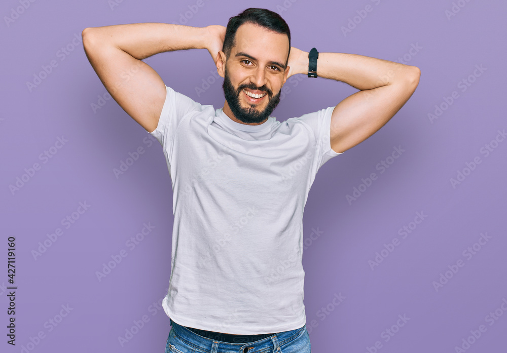 Young man with beard wearing casual white t shirt relaxing and stretching, arms and hands behind head and neck smiling happy Young man with beard wearing casual white t shirt relaxing and stretching, arms and hands behind head and neck smiling happy