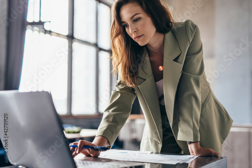 Woman stands leaning on her desk and uses her laptop