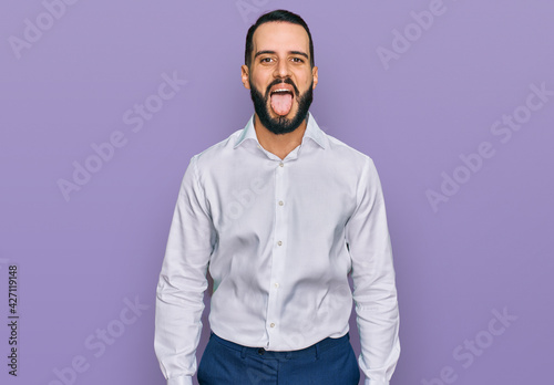 Young man with beard wearing business shirt sticking tongue out happy with funny expression. emotion concept.