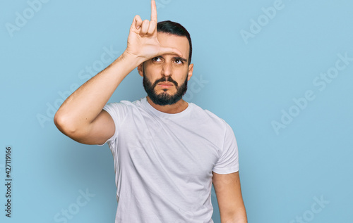 Young man with beard wearing casual white t shirt making fun of people with fingers on forehead doing loser gesture mocking and insulting.
