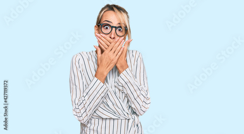 Beautiful blonde woman wearing business shirt and glasses shocked covering mouth with hands for mistake. secret concept.