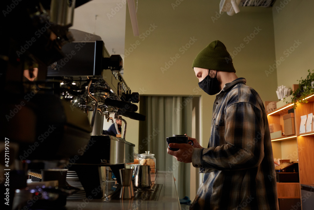 Side view portrait of male barista wearing mask while making fresh coffee at bar counter in cafe , copy space Side view portrait of male barista wearing mask while making fresh coffee at bar counter in cafe , copy space
