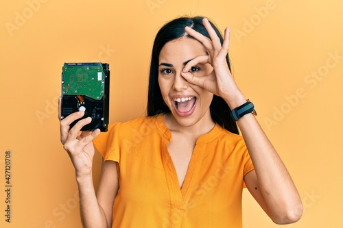 Beautiful young woman holding hard disk smiling happy doing ok sign with hand on eye looking through fingers