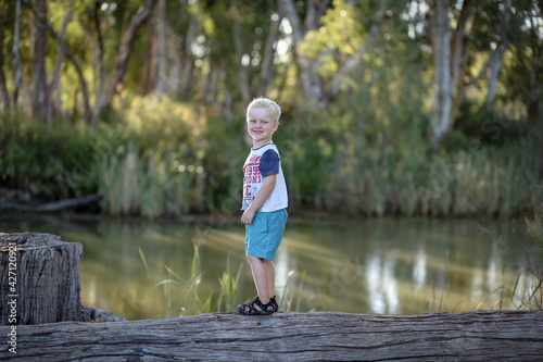 Little blonde caucasian boy enjoying outdoor adventure by the river climbing on old logs