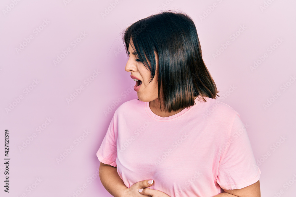 Young hispanic woman wearing casual pink t shirt with hand on stomach because nausea, painful disease feeling unwell. ache concept. Young hispanic woman wearing casual pink t shirt with hand on stomach because nausea, painful disease feeling unwell. ache concept.