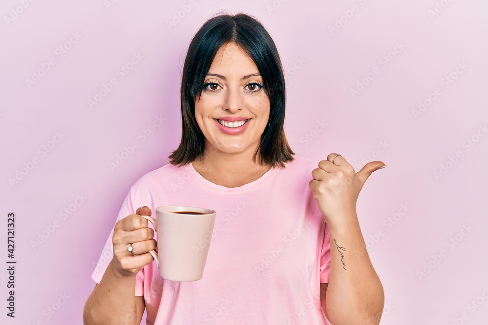 Young hispanic woman drinking a cup of coffee pointing thumb up to the side smiling happy with open mouth Young hispanic woman drinking a cup of coffee pointing thumb up to the side smiling happy with open mouth