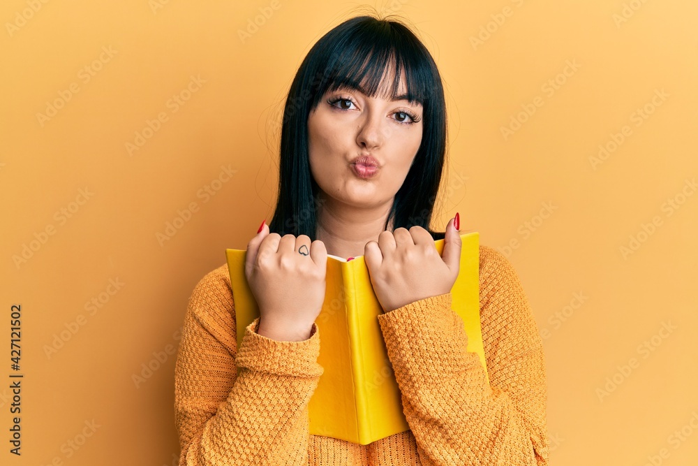 Young hispanic woman holding book looking at the camera blowing a kiss being lovely and sexy. love expression. Young hispanic woman holding book looking at the camera blowing a kiss being lovely and sexy. love expression.