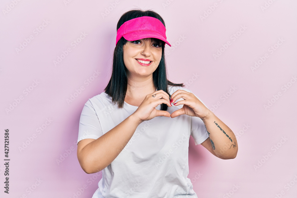 Young hispanic woman wearing sportswear and sun visor cap smiling in love showing heart symbol and shape with hands. romantic concept.
