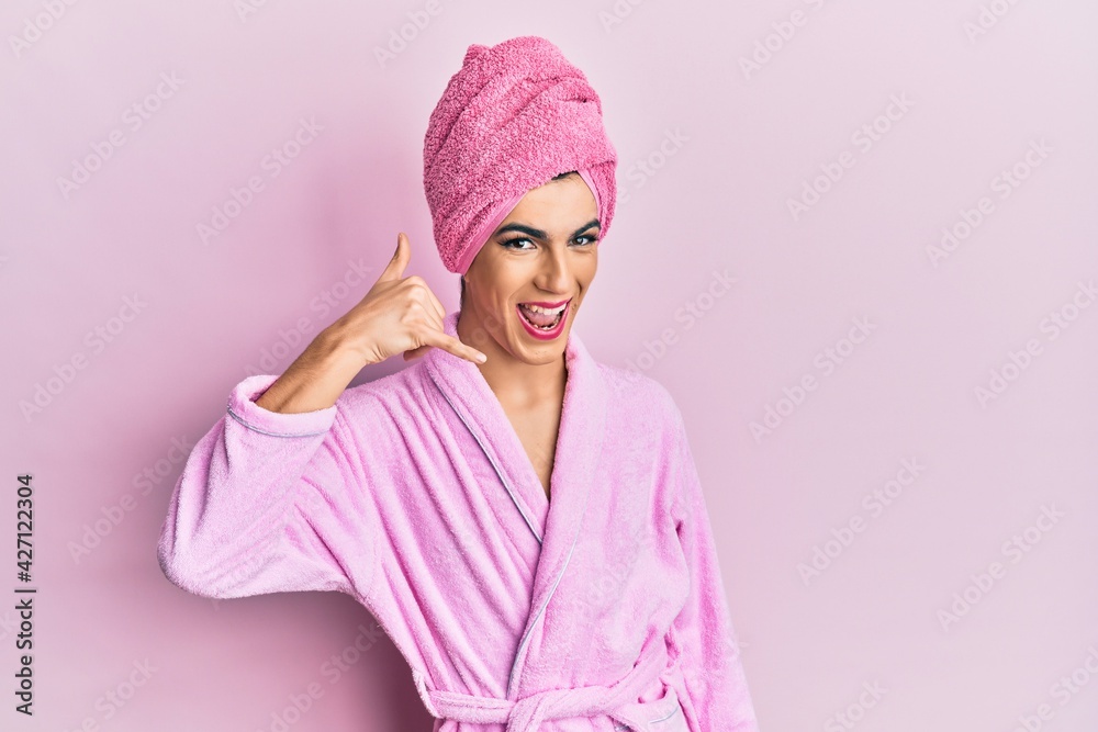Young man wearing woman make up wearing shower towel on head and bathrobe smiling doing phone gesture with hand and fingers like talking on the telephone. communicating concepts. Young man wearing woman make up wearing shower towel on head and bathrobe smiling doing phone gesture with hand and fingers like talking on the telephone. communicating concepts.