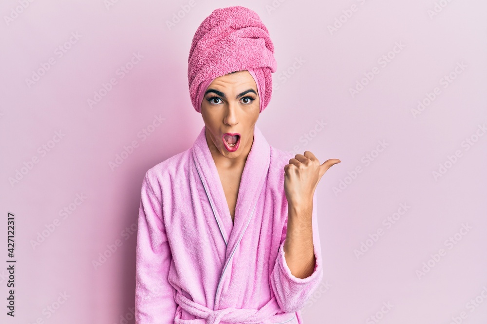 Young man wearing woman make up wearing shower towel on head and bathrobe surprised pointing with hand finger to the side, open mouth amazed expression. Young man wearing woman make up wearing shower towel on head and bathrobe surprised pointing with hand finger to the side, open mouth amazed expression.