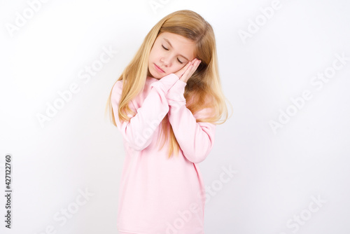 Relax and sleep time. Tired beautiful caucasian little girl wearing pink hoodie over white background with closed eyes leaning on palms making sleeping gesture.