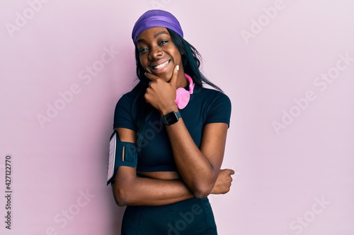 Young african american woman wearing gym clothes and using headphones looking confident at the camera smiling with crossed arms and hand raised on chin. thinking positive.