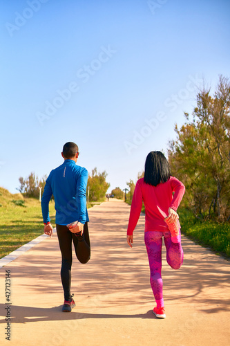 Couple stretching their legs before training. The woman wears bright pink and purple clothes. Man is wearing blue shirt and black long trousers. Wearing sunglasses. Shot from behind.