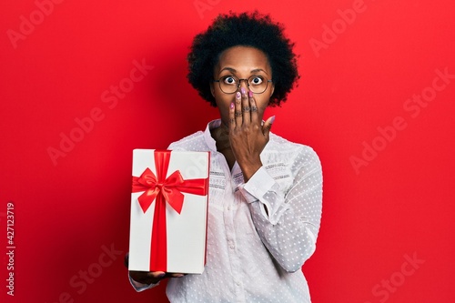 Young african american woman holding gift covering mouth with hand, shocked and afraid for mistake. surprised expression © Krakenimages.com