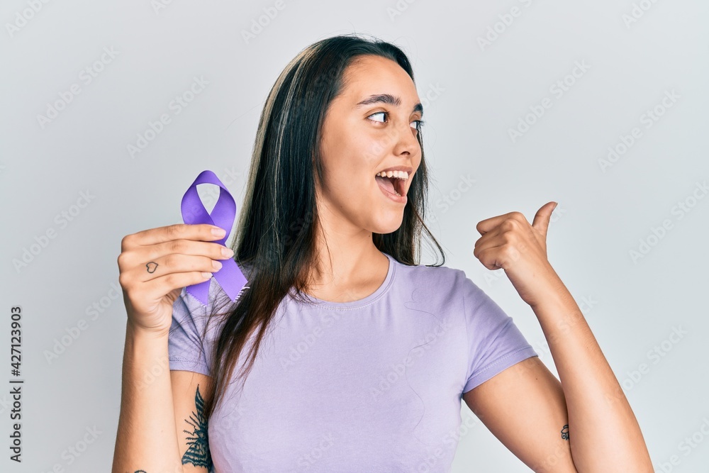 Young hispanic woman holding purple ribbon awareness pointing thumb up to the side smiling happy with open mouth Young hispanic woman holding purple ribbon awareness pointing thumb up to the side smiling happy with open mouth