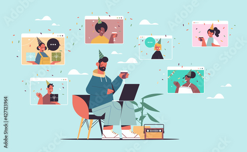 man in festive hat using laptop celebrating online birthday party with mix race friends in computer windows