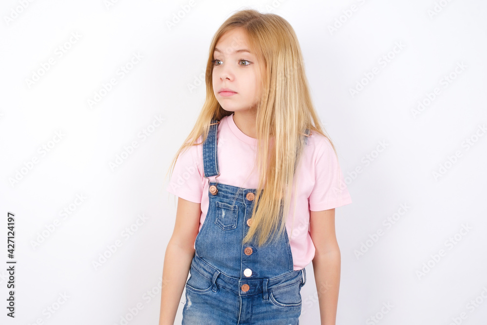 beautiful caucasian little girl wearing jeans overall over white background stares aside with wondered expression has speechless expression. Embarrassed model looks in surprise beautiful caucasian little girl wearing jeans overall over white background stares aside with wondered expression has speechless expression. Embarrassed model looks in surprise