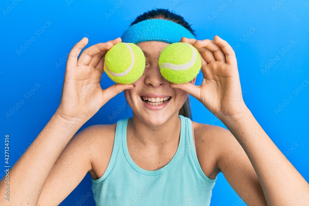 Beautiful brunette little girl holding tennis ball close to eyes smiling  and laughing hard out loud because funny crazy joke. Photos | Adobe Stock