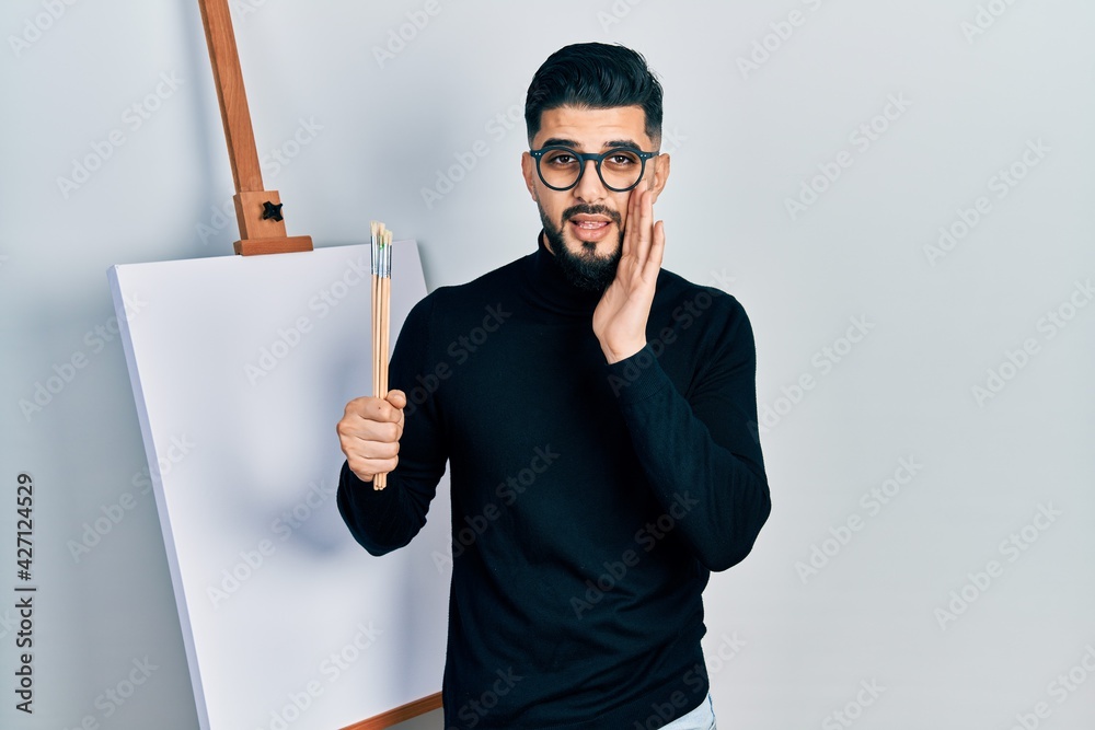 Handsome man with beard holding brushes close to easel stand hand on mouth telling secret rumor, whispering malicious talk conversation Handsome man with beard holding brushes close to easel stand hand on mouth telling secret rumor, whispering malicious talk conversation
