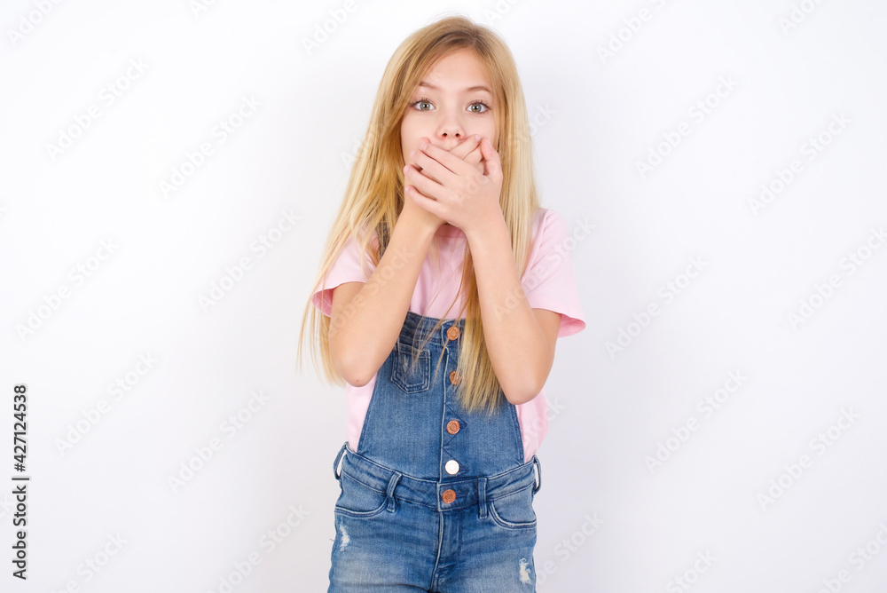 Stunned beautiful caucasian little girl wearing jeans overall over white background covers mouth with both hands being afraid from something or after hearing stunning gossips. Stunned beautiful caucasian little girl wearing jeans overall over white background covers mouth with both hands being afraid from something or after hearing stunning gossips.