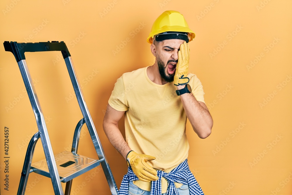 Handsome man with beard by construction stairs wearing hardhat yawning tired covering half face, eye and mouth with hand. face hurts in pain. Handsome man with beard by construction stairs wearing hardhat yawning tired covering half face, eye and mouth with hand. face hurts in pain.