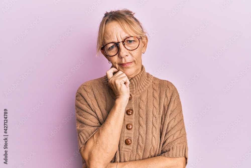 Middle age blonde woman wearing glasses over pink background thinking concentrated about doubt with finger on chin and looking up wondering