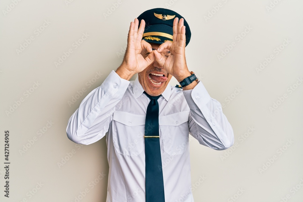 Handsome middle age mature man wearing airplane pilot uniform doing ok gesture like binoculars sticking tongue out, eyes looking through fingers. crazy expression. Handsome middle age mature man wearing airplane pilot uniform doing ok gesture like binoculars sticking tongue out, eyes looking through fingers. crazy expression.