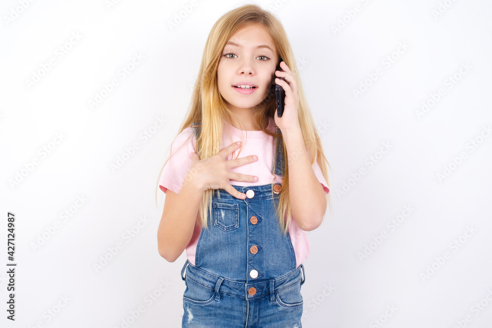 Smiling beautiful caucasian little girl wearing jeans overall over white background talks via cellphone, enjoys pleasant great conversation. People, technology, communication concept Smiling beautiful caucasian little girl wearing jeans overall over white background talks via cellphone, enjoys pleasant great conversation. People, technology, communication concept
