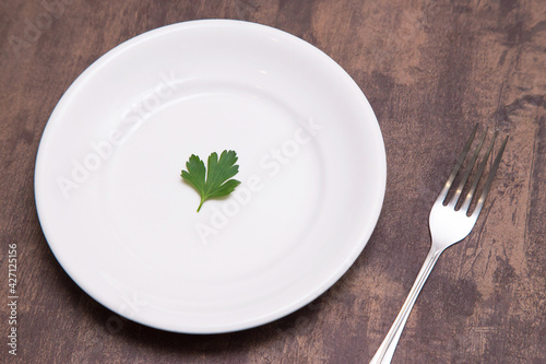 Single one parsley green leaf on white empty plate and fork on table. Diet, fasting, vegan, vegetarian, healthy food concept
