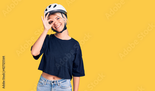 Young beautiful blonde woman wearing bike helmet smiling happy doing ok sign with hand on eye looking through fingers