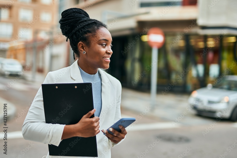 Young african american businesswoman using smartphone holding clipboard at the city. Young african american businesswoman using smartphone holding clipboard at the city.