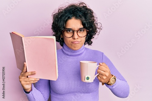 Young hispanic woman with curly hair reading a book and drinking a cup of coffee skeptic and nervous, frowning upset because of problem. negative person.