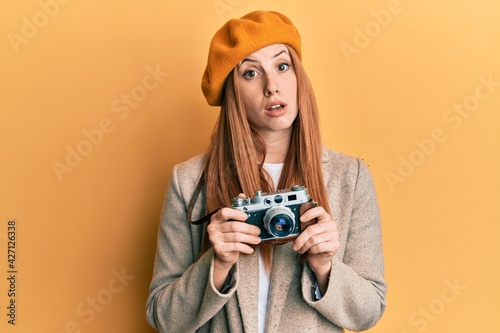 Young irish woman holding vintage camera in shock face, looking skeptical and sarcastic, surprised with open mouth