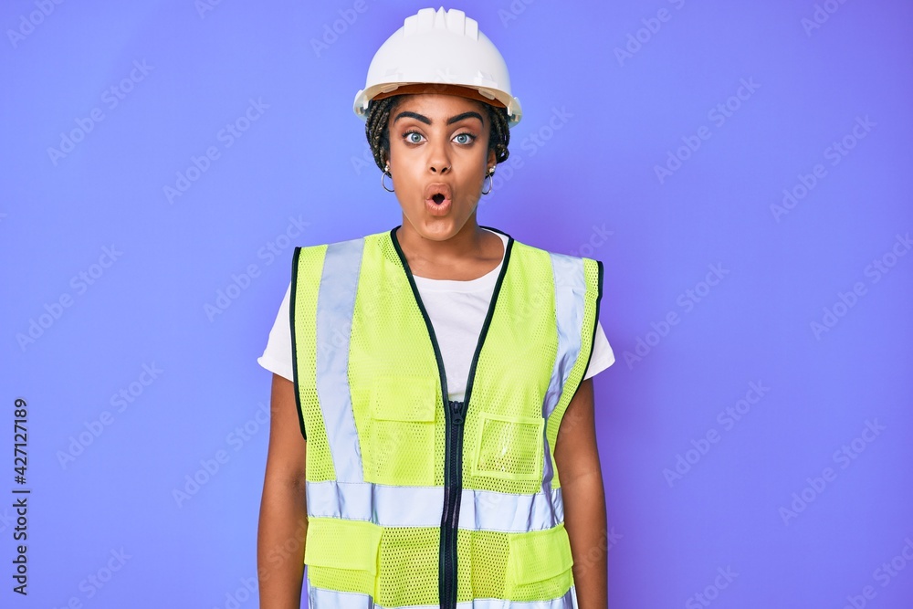 Young african american woman with braids wearing safety helmet and reflective jacket scared and amazed with open mouth for surprise, disbelief face
