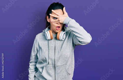 Young hispanic woman wearing gym clothes and using headphones peeking in shock covering face and eyes with hand, looking through fingers afraid © Krakenimages.com