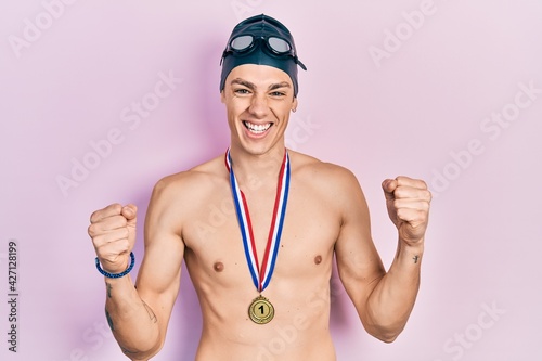 Young hispanic man wearing swimmer glasses and gold medal screaming proud, celebrating victory and success very excited with raised arms © Krakenimages.com