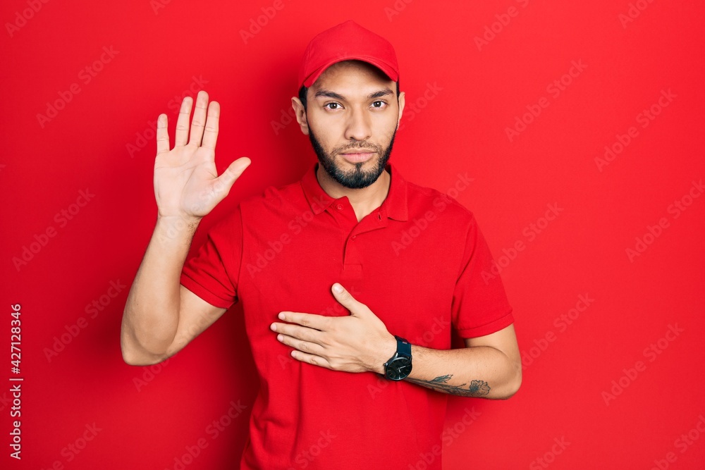 Hispanic man with beard wearing delivery uniform and cap swearing with hand on chest and open palm, making a loyalty promise oath Hispanic man with beard wearing delivery uniform and cap swearing with hand on chest and open palm, making a loyalty promise oath