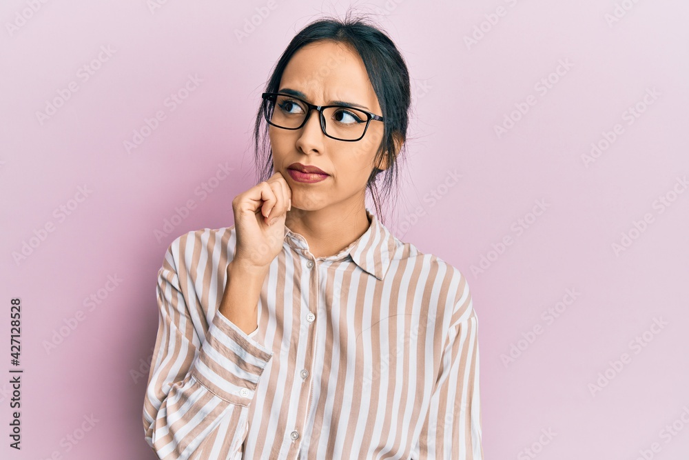 Young hispanic girl wearing casual clothes and glasses thinking concentrated about doubt with finger on chin and looking up wondering