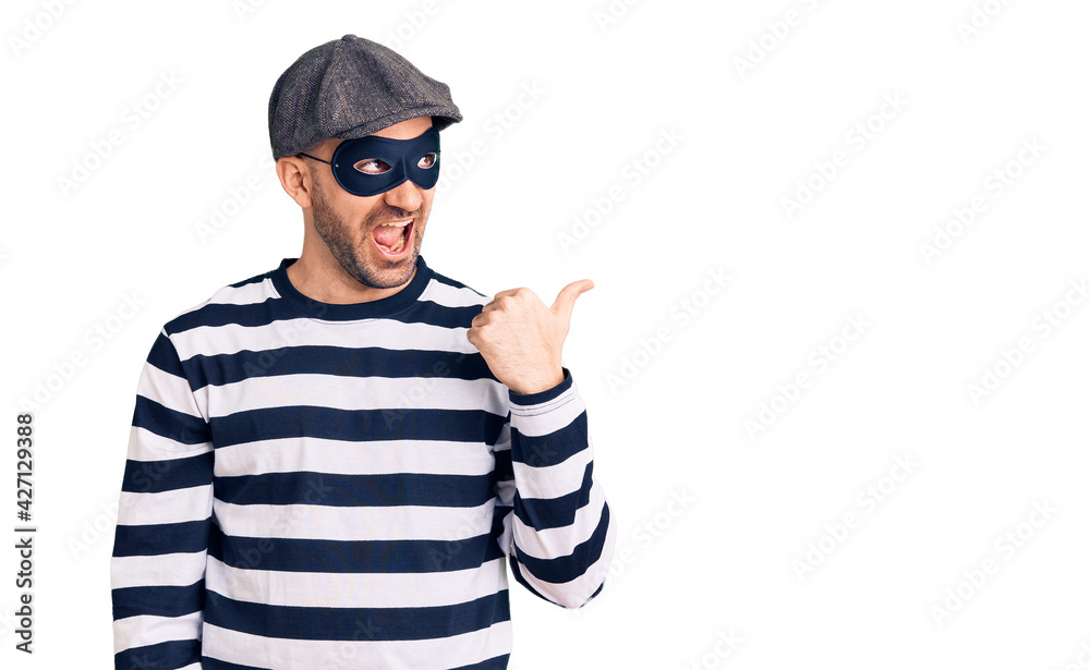 Young handsome man wearing burglar mask smiling with happy face looking and pointing to the side with thumb up. Young handsome man wearing burglar mask smiling with happy face looking and pointing to the side with thumb up.