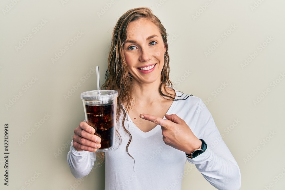 Young blonde woman drinking glass of soda beverage smiling happy pointing with hand and finger Young blonde woman drinking glass of soda beverage smiling happy pointing with hand and finger