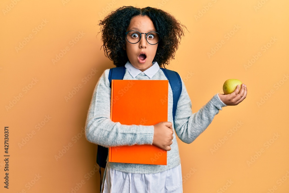 Young little girl with afro hair wearing school bag holding books and green apple afraid and shocked with surprise and amazed expression, fear and excited face. Young little girl with afro hair wearing school bag holding books and green apple afraid and shocked with surprise and amazed expression, fear and excited face.