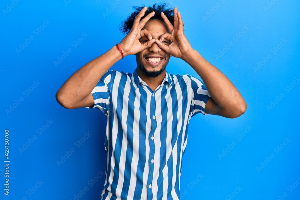 Young african american man with beard wearing casual striped shirt doing ok gesture like binoculars sticking tongue out, eyes looking through fingers. crazy expression. Young african american man with beard wearing casual striped shirt doing ok gesture like binoculars sticking tongue out, eyes looking through fingers. crazy expression.